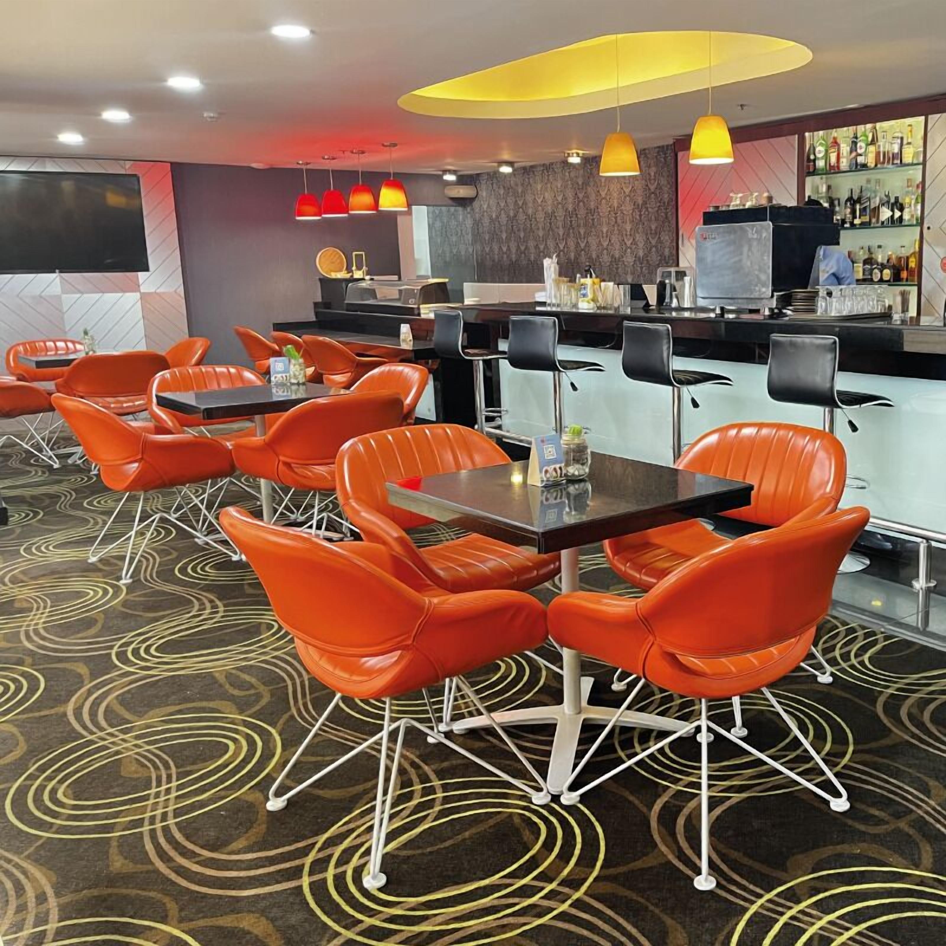 Um restaurante com cadeiras e mesas laranja, área de bar e carpete estampado.