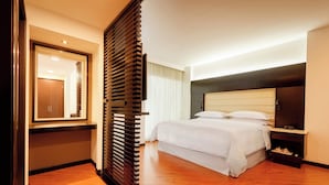 A modern hotel room with a wooden floor, a large bed with white linens, a nightstand with a lamp, and a large mirror on the wall.