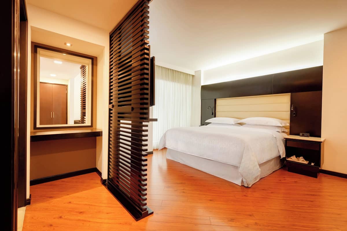 A modern hotel room with a wooden floor, a large bed with white linens, a nightstand with a lamp, and a large mirror on the wall.