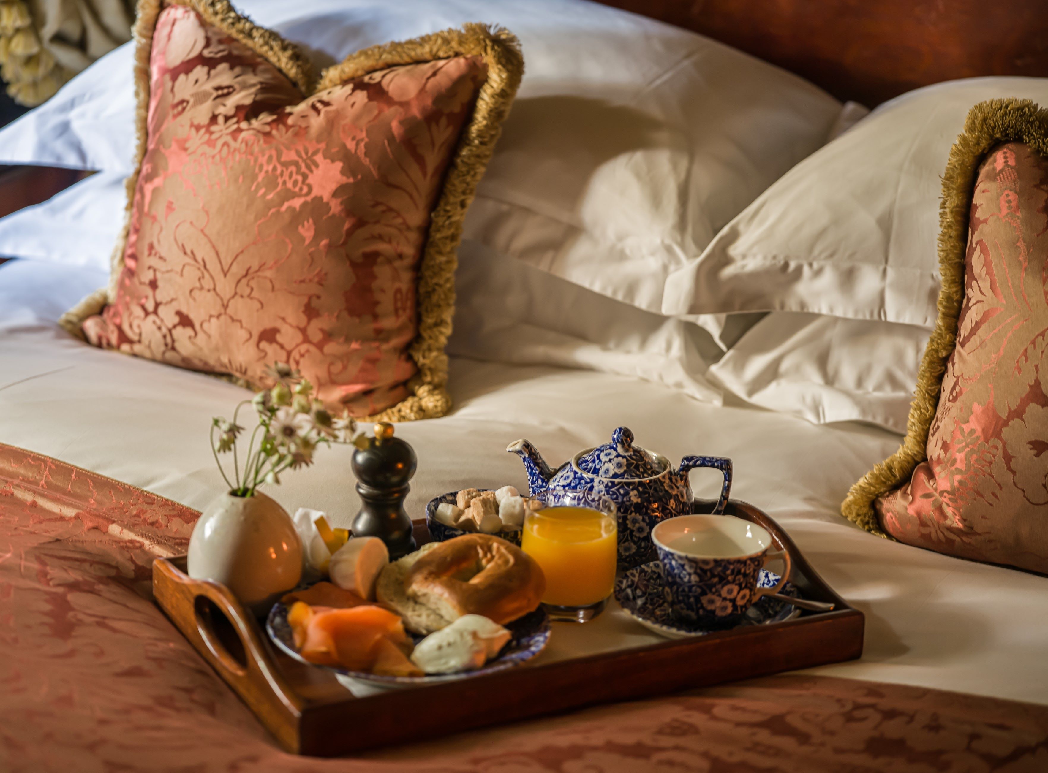 A neatly made bed with a tray of breakfast items including eggs, toast, and orange juice, set against a backdrop of patterned pillows and a wooden headboard.