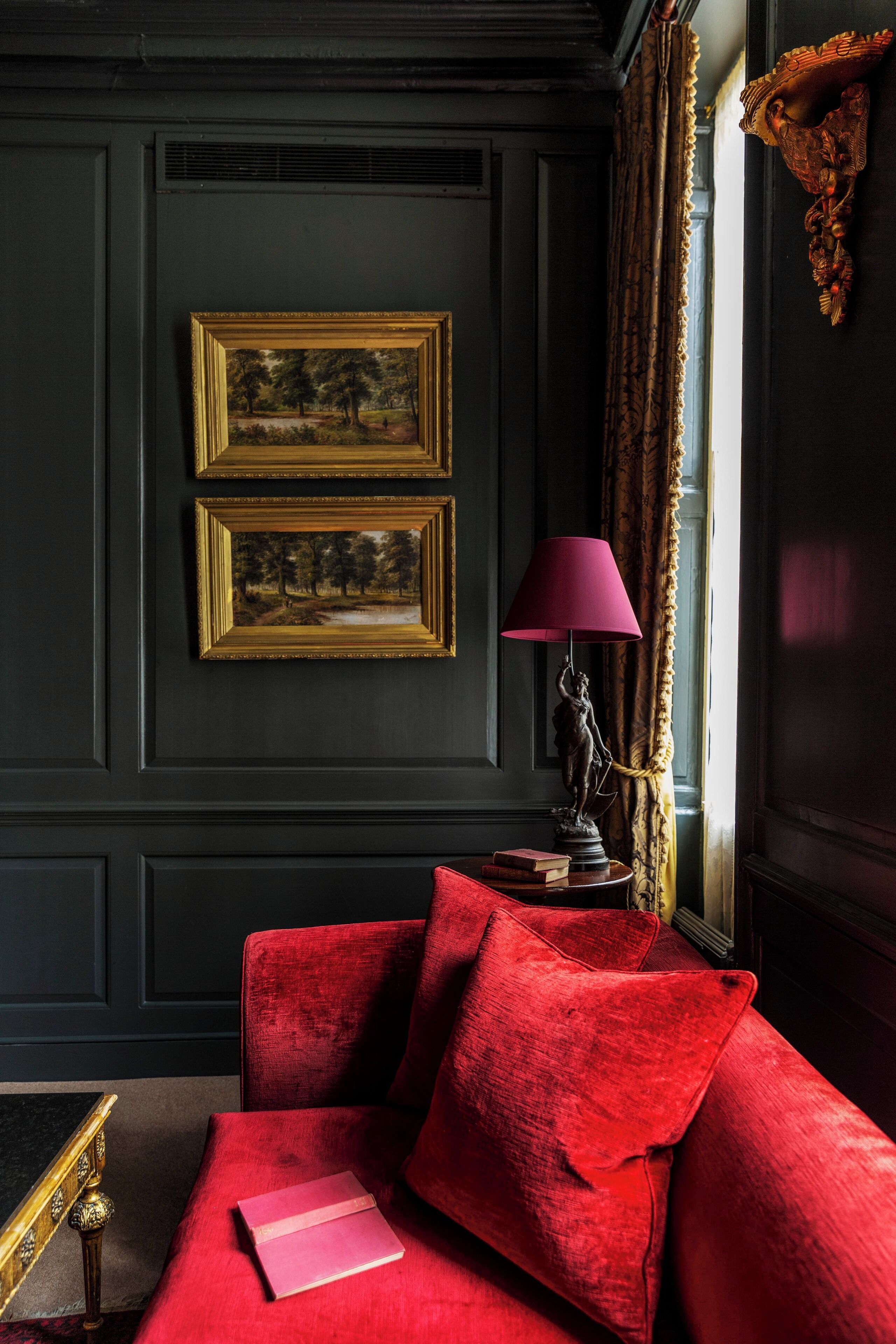 A dark room with a red sofa, two framed paintings, a statue, and a lamp.