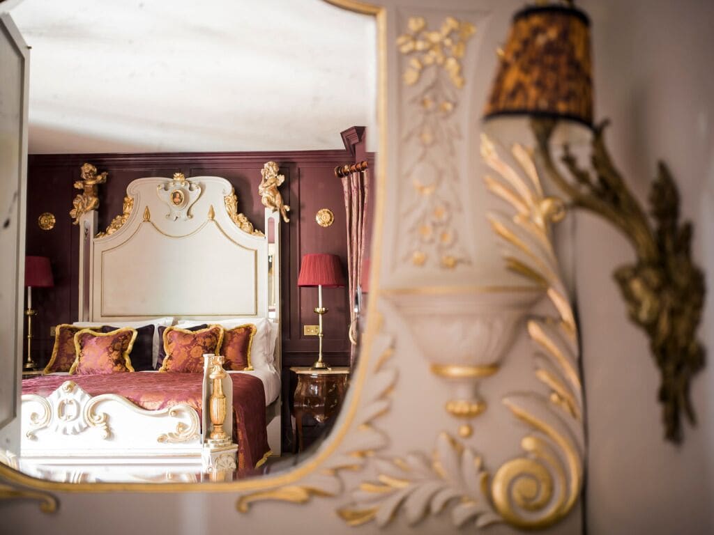 A luxurious bedroom with an ornate bed, a mirror reflecting the room, and a decorative lamp.