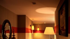 A hotel hallway with a red carpet, a wooden console table with a mirror, a wall-mounted lamp, and a painting on the wall. - Le Boutique Hôtel Garonne by Occitania hotels (Toulouse)