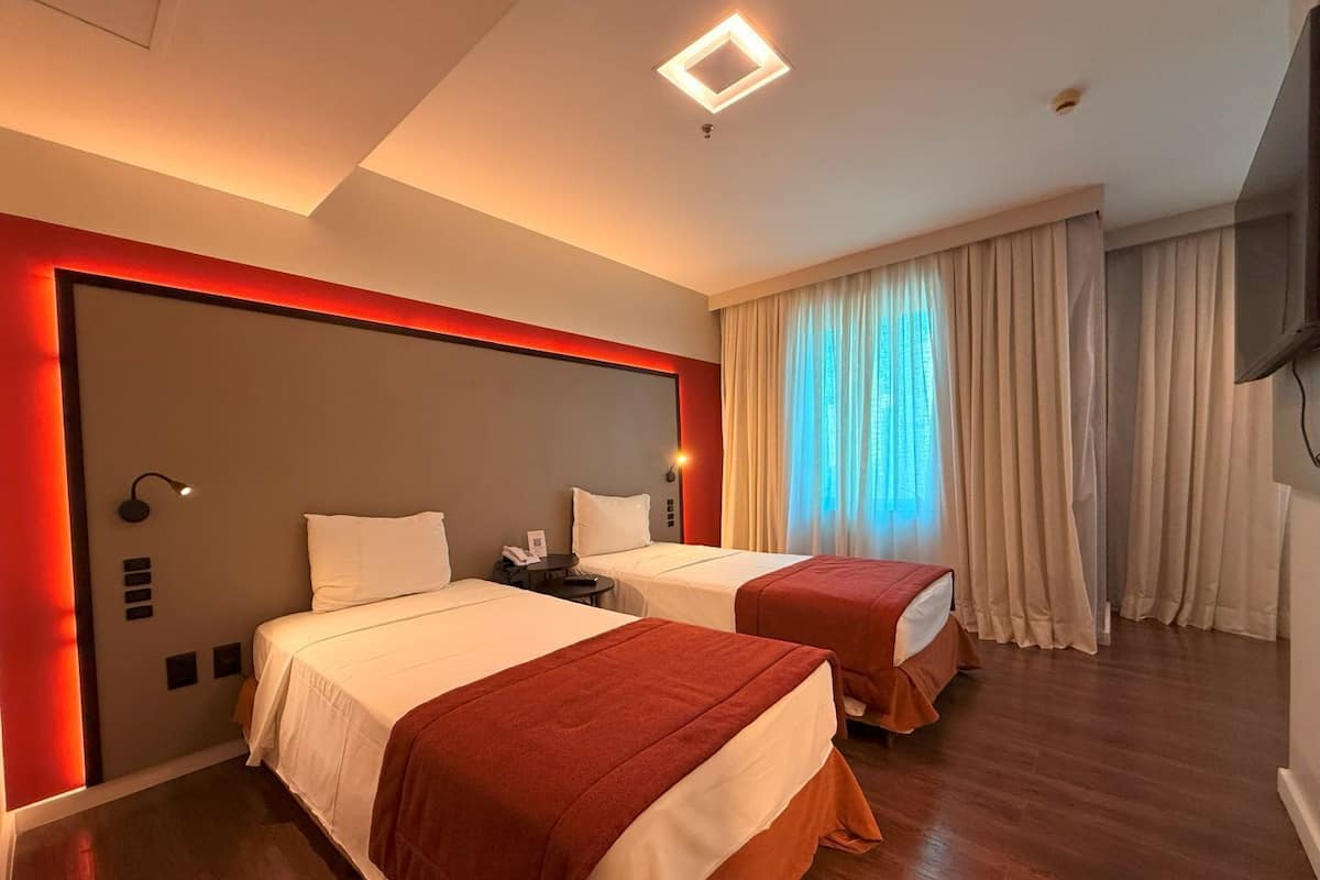A hotel room with two beds, a red accent wall, a ceiling light, a TV, and a window with curtains.