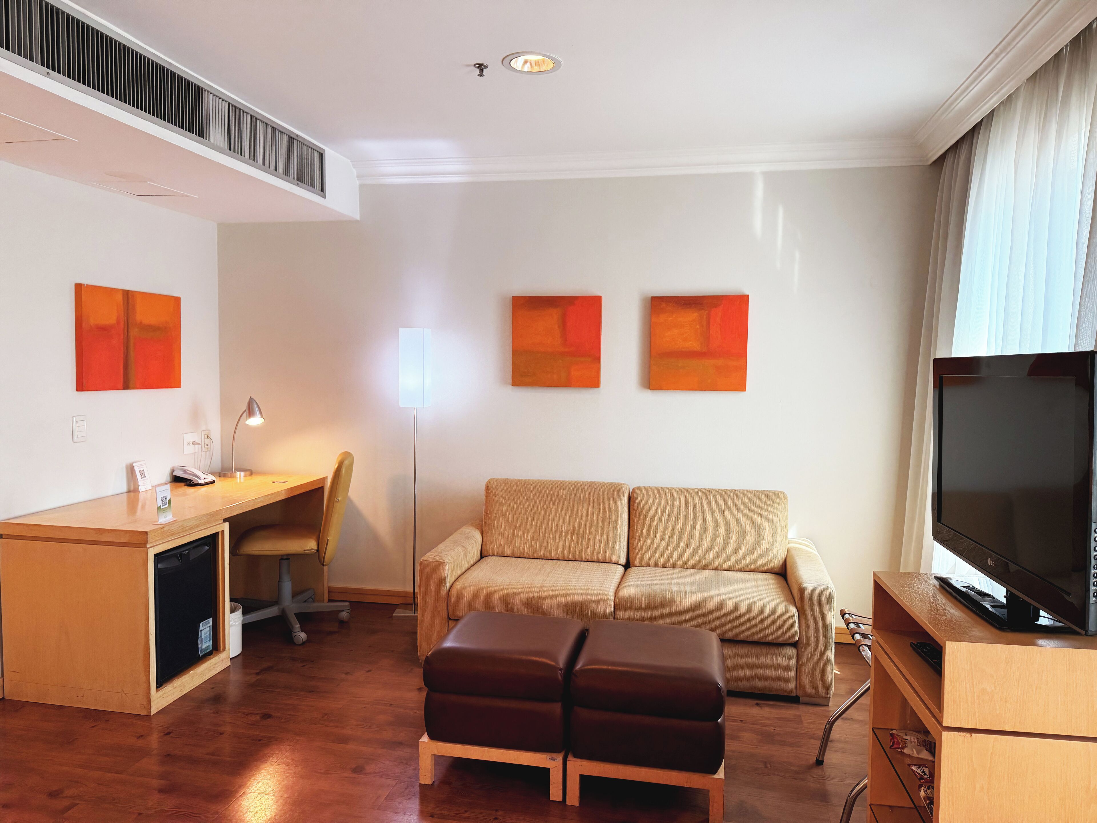 A modern hotel room with a wooden desk, a sofa, a TV, and artwork on the walls.