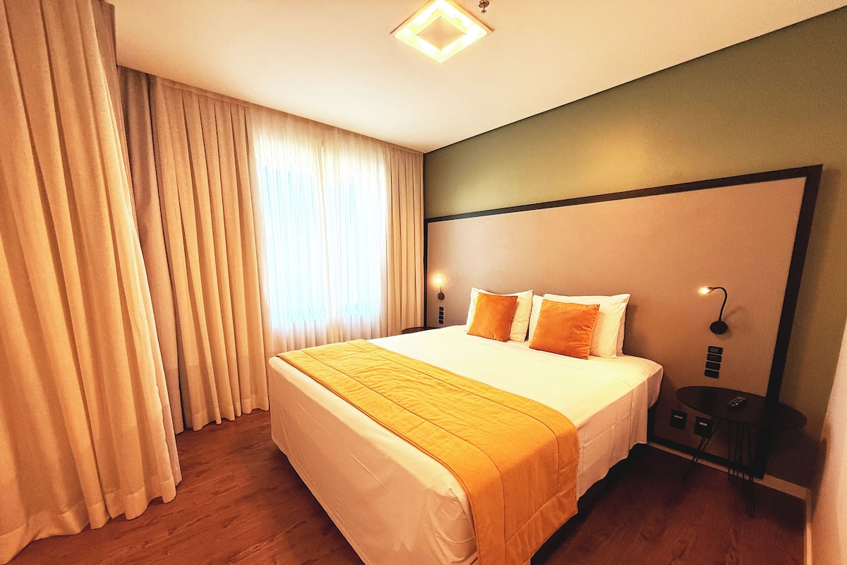 A hotel room with a large bed, a bedside table, a nightstand, and a wall-mounted lamp.
