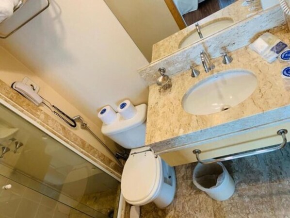 A bathroom with a sink, mirror, and toilet. There is a hairdryer on the wall and a trash bin next to the toilet.
