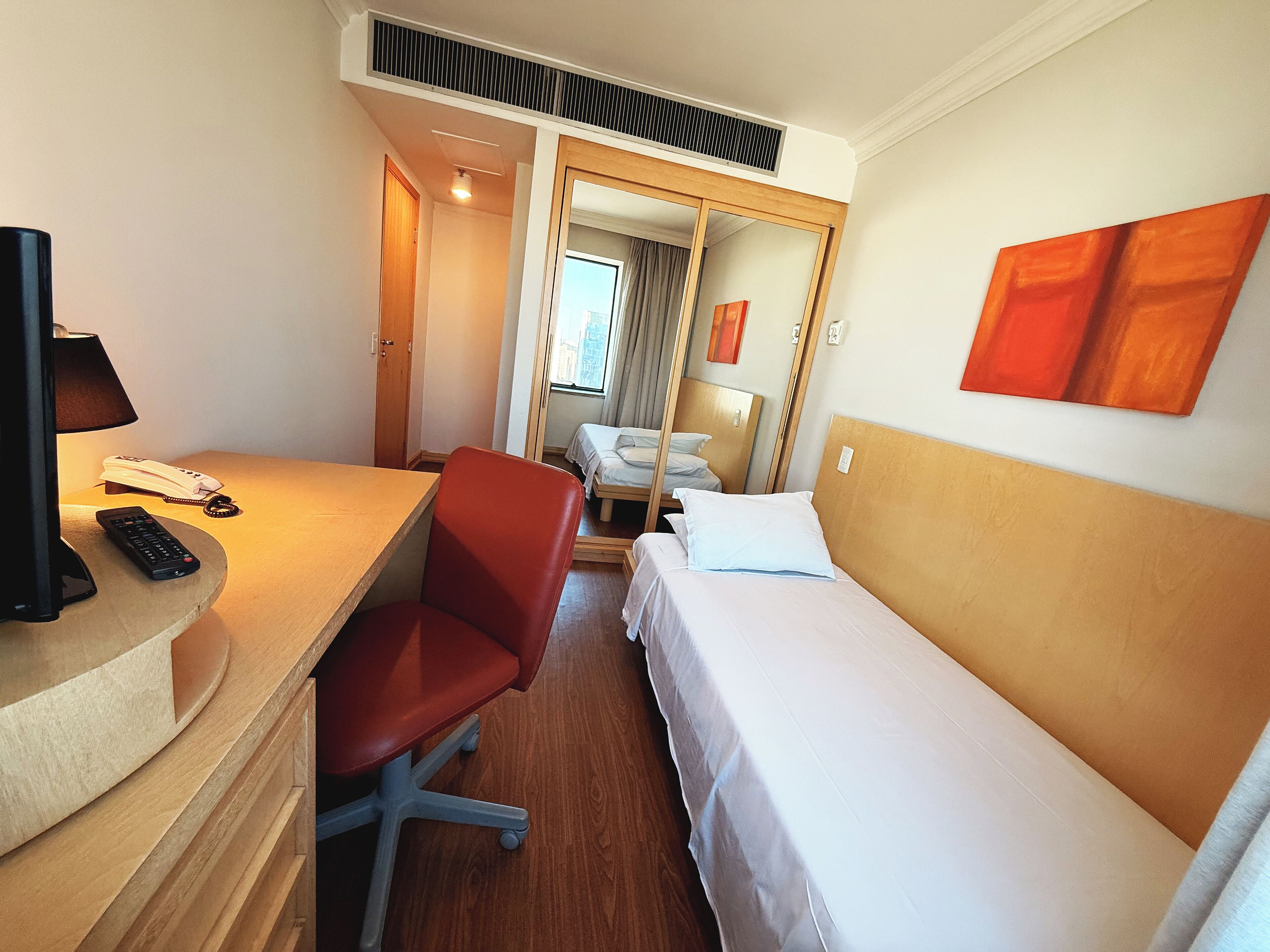 A hotel room with a desk, a red office chair, a bed, and a large mirror.