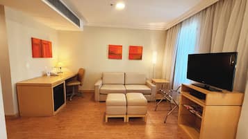 Quarto de hotel moderno com piso de madeira, um sofá, uma escrivaninha com luminária, uma televisão e duas pinturas na parede.