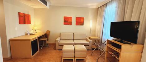 Una habitación de hotel moderna con piso de madera, un sofá, un escritorio con lámpara, un televisor y dos cuadros en la pared.