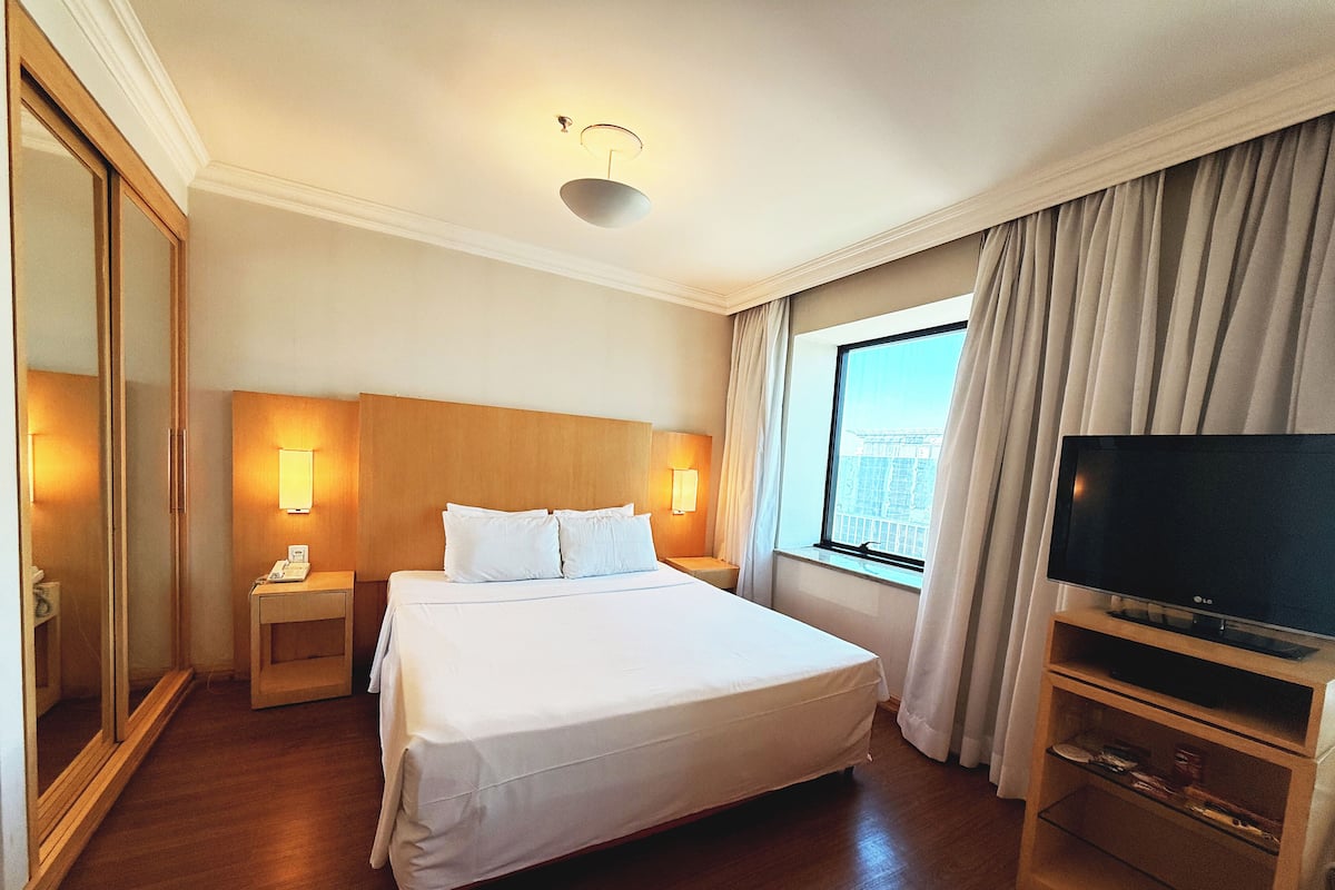A hotel room with a large bed, bedside lamps, a TV on a wooden stand, and a window with curtains.