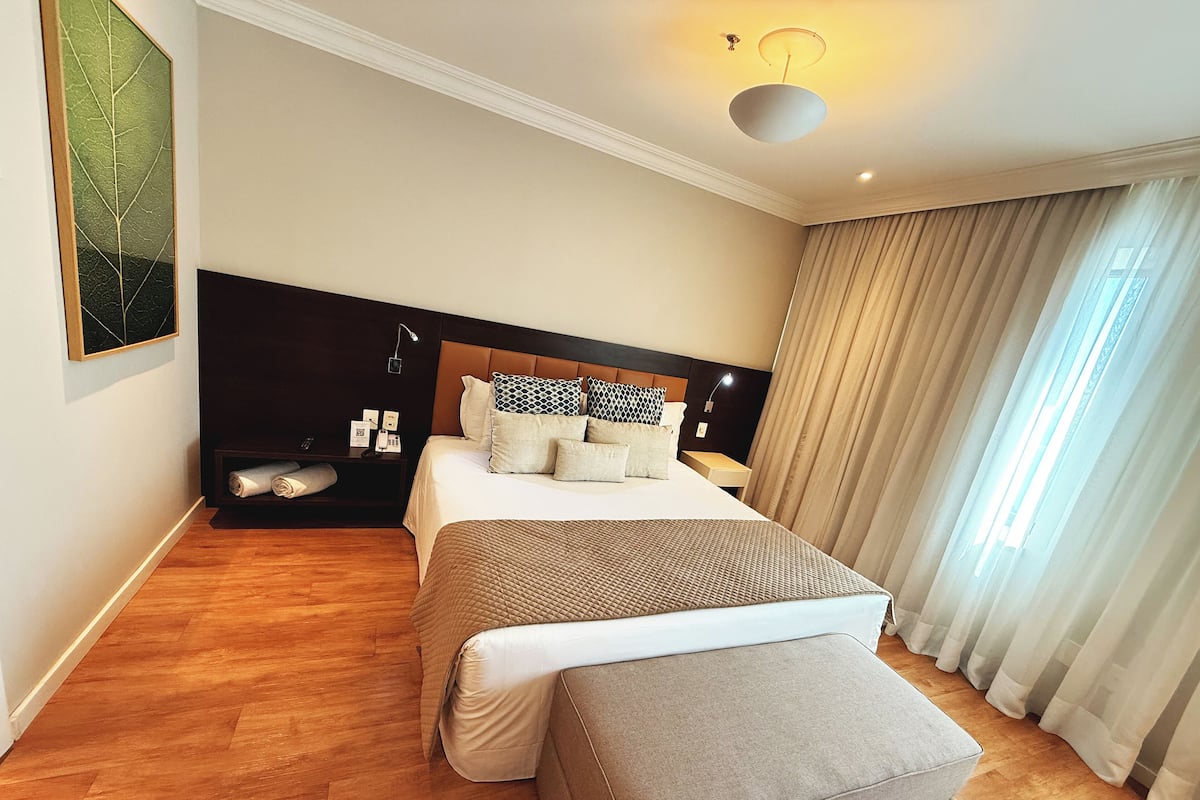 A modern hotel room with a large bed, a bedside table with a lamp, a nightstand, a wall-mounted phone, and a large window with curtains.