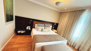 A modern hotel room with a large bed, a bedside table with a lamp, a nightstand, a wall-mounted phone, and a large window with curtains.