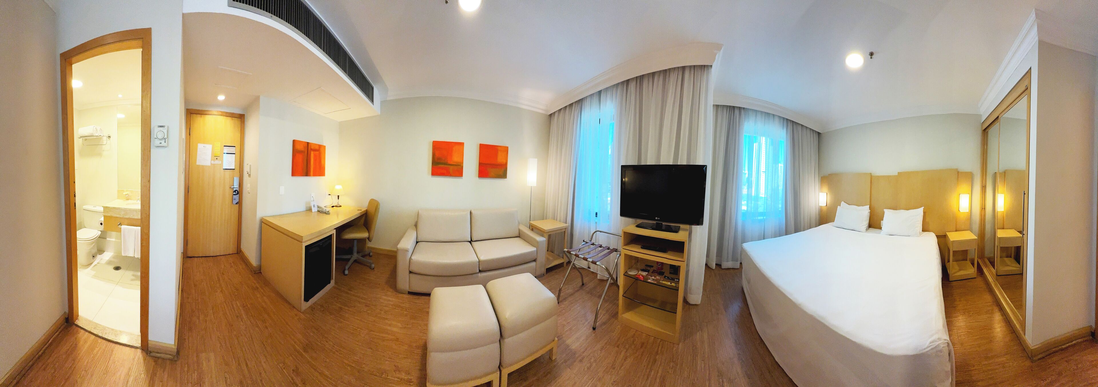 A hotel room with a bed, a desk, a sofa, a TV, and a bathroom visible through an open door.