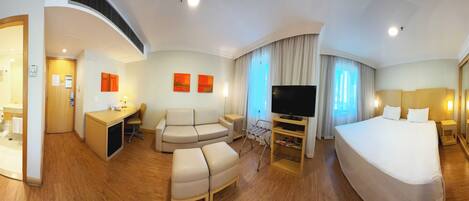 A hotel room with a bed, a desk, a sofa, a TV, and a bathroom visible through an open door.
