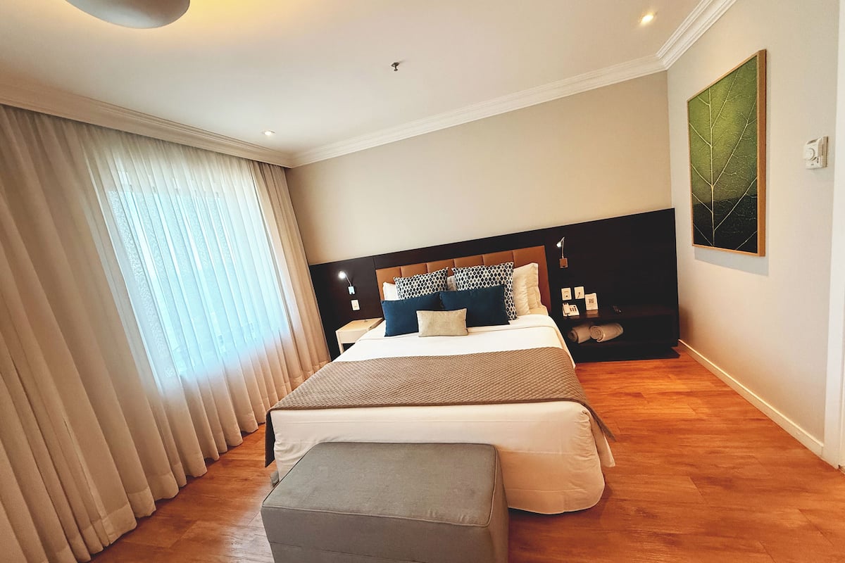 A modern bedroom with a large bed, a bedside table, a nightstand, a bench, and a window with curtains.