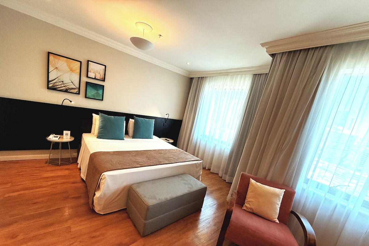 A hotel room with a bed, a small table with a phone, a chair, and a large window with curtains.