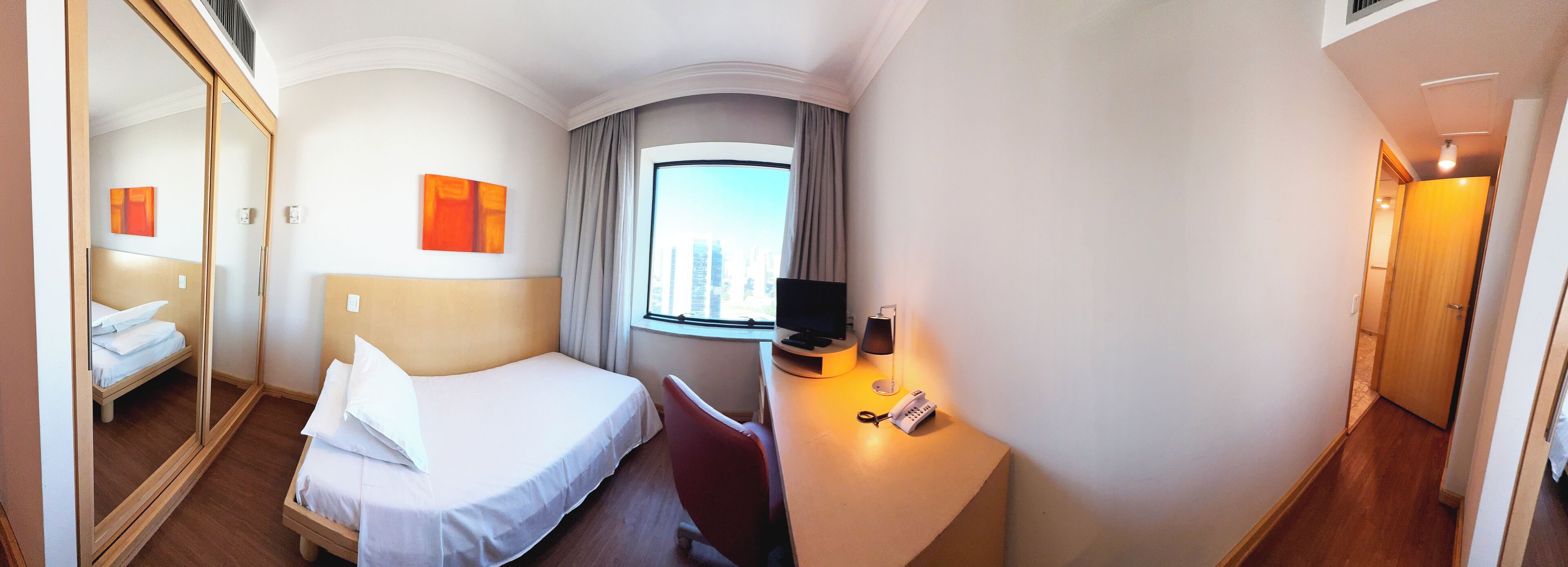 Habitación de hotel con cama, escritorio con lámpara, silla, ventana con cortinas y vista a la ciudad.