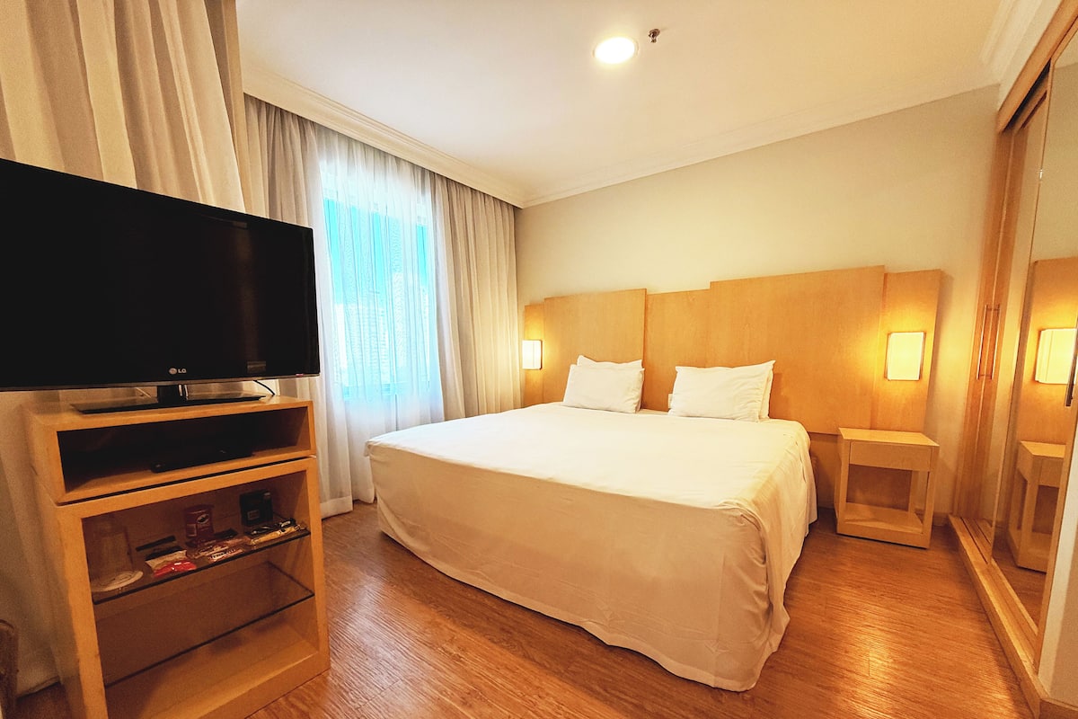 A hotel room with a large bed, a TV on a wooden stand, and a small table with a lamp.