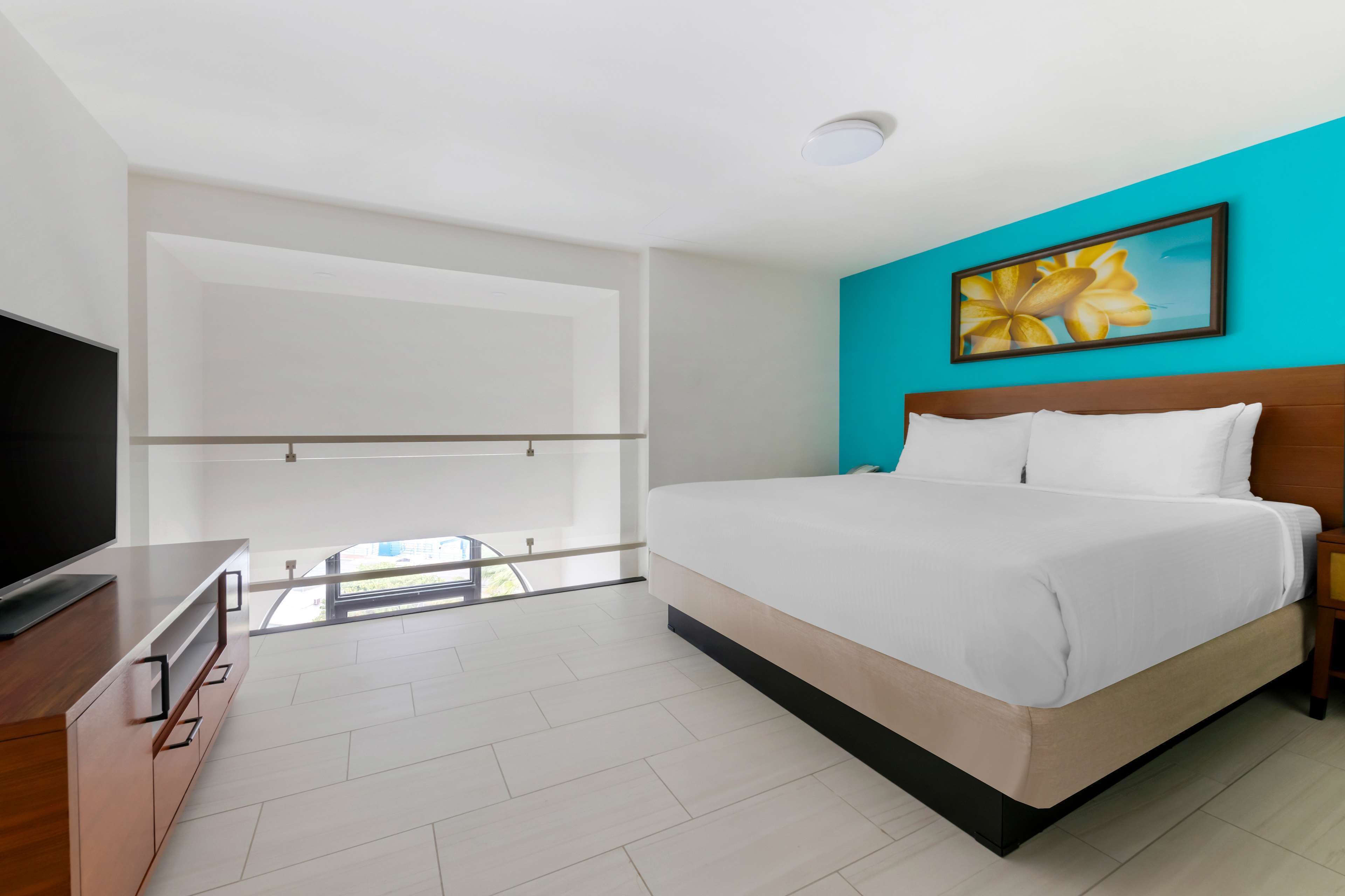 A modern hotel room with a large bed, a television on a wooden dresser, and a balcony with a glass door.
