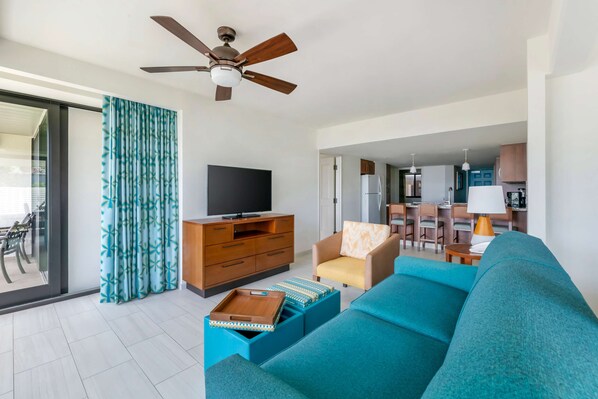 A modern living room with a sofa, a television, and a ceiling fan. There is a kitchen area visible in the background.