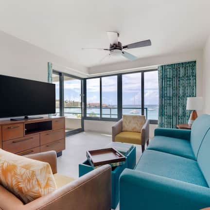 A modern living room with a flat-screen TV, a ceiling fan, and a large window offering a view of the sea.