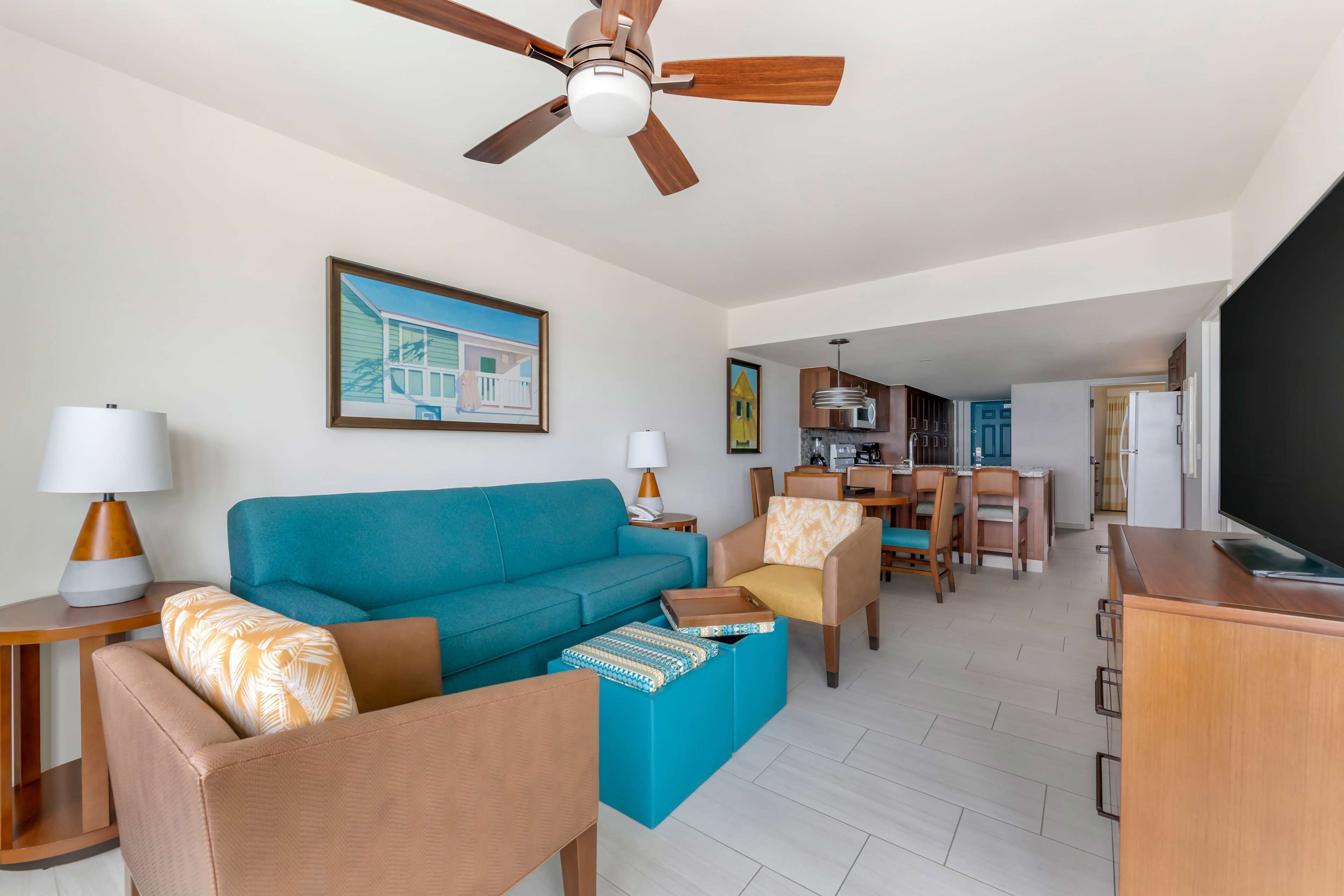 A living room with a teal sofa, a wooden coffee table, and a ceiling fan. There is a dining area with chairs and a table, and a kitchen in the background.
