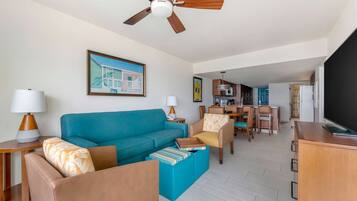 A living room with a teal sofa, a wooden coffee table, and a ceiling fan. There is a dining area with chairs and a table, and a kitchen in the background.