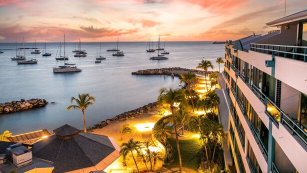 Een resort met uitzicht op zeilboten op het water, een strand met palmbomen en een modern gebouw met balkons.