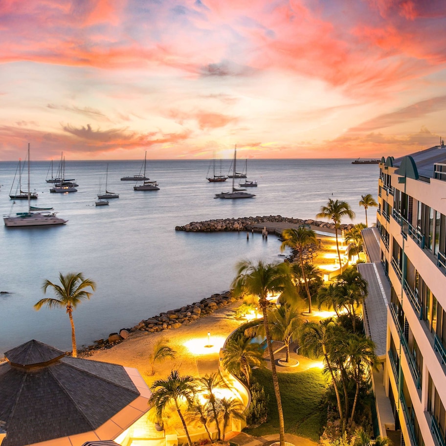 A resort with a view of sailboats on the water, a beach with palm trees, and a modern building with balconies.