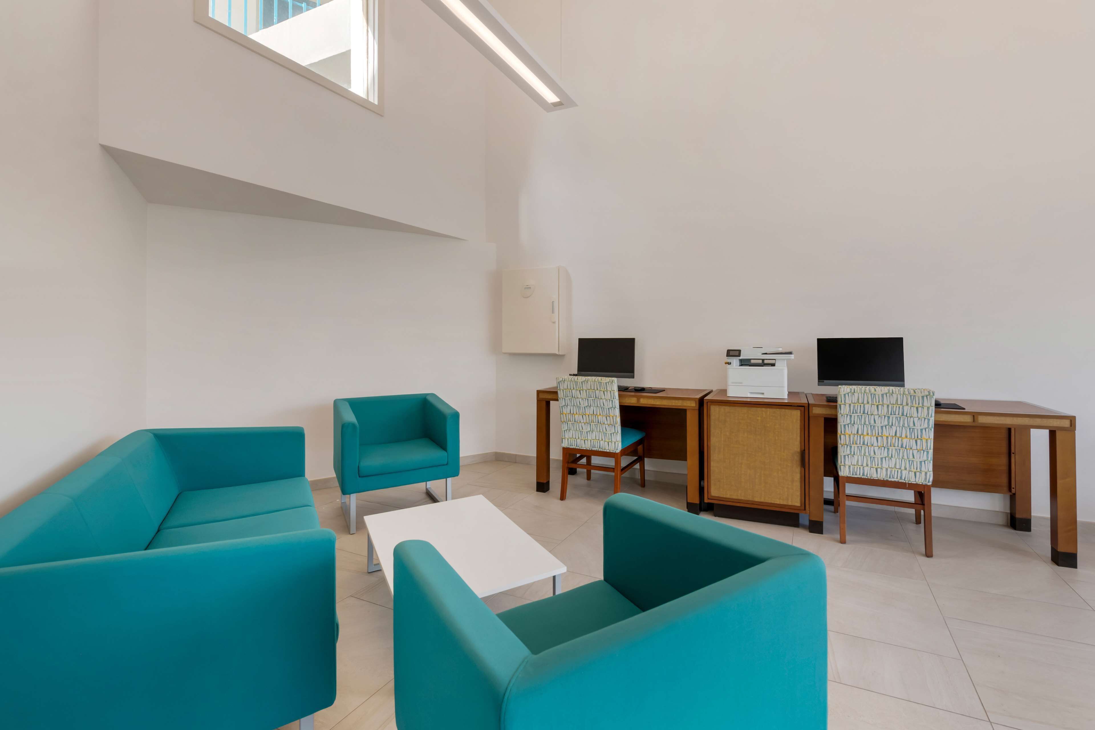 A modern room with teal sofas, a wooden desk, two monitors, and a white cabinet.