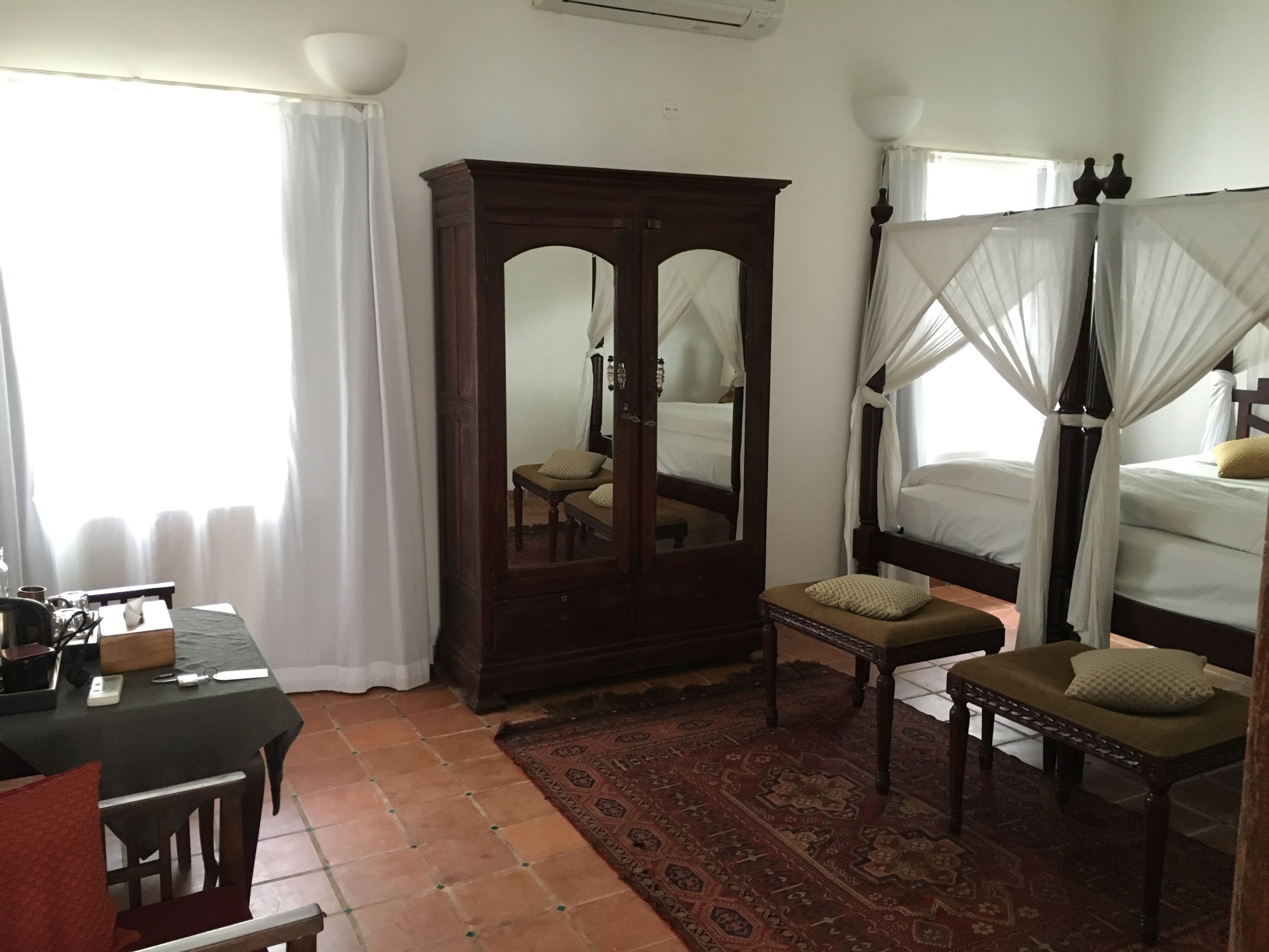 A bedroom with a large wooden wardrobe, a bed with a canopy, a small table with a chair, and a red patterned rug.