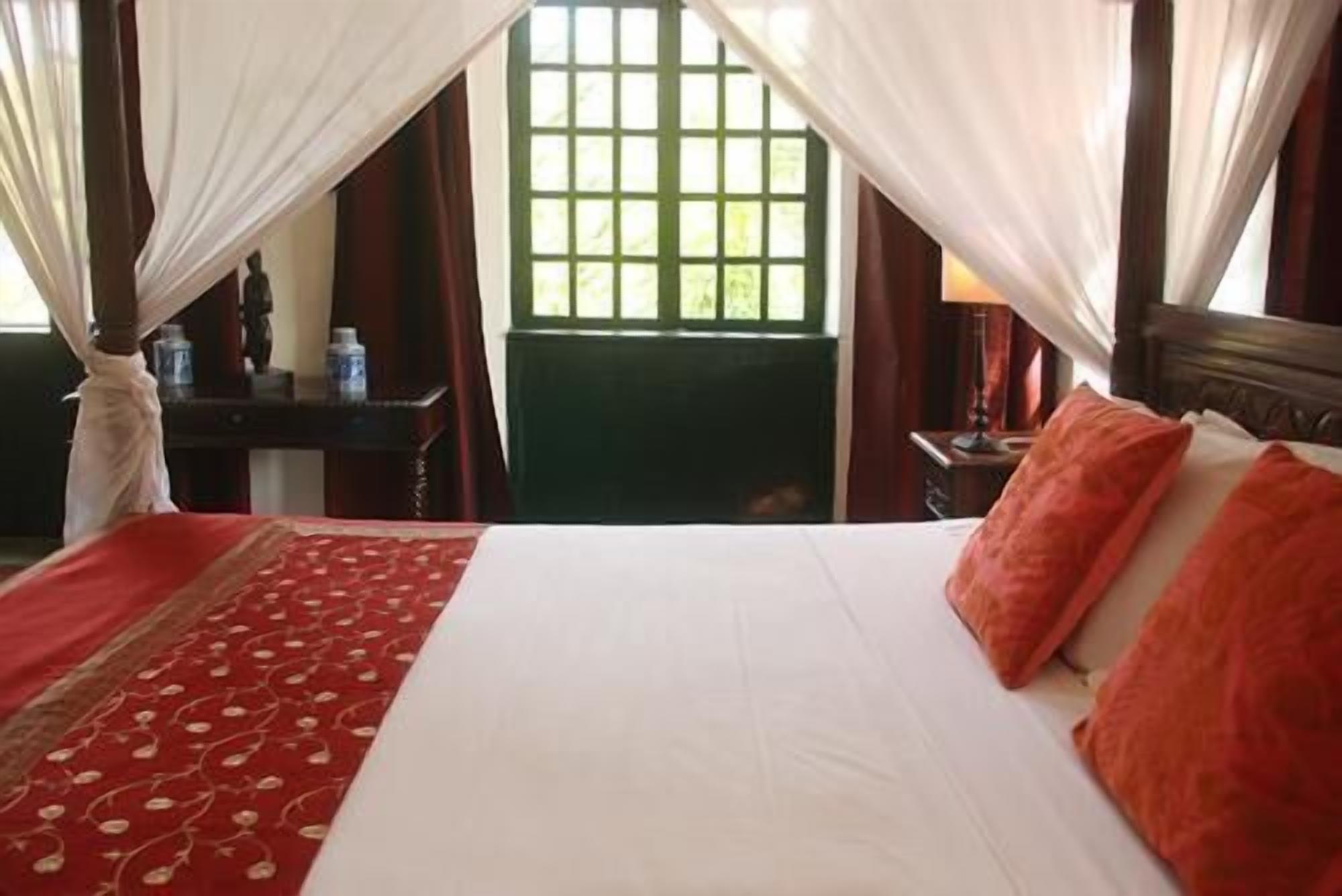 Una habitación de hotel con una cama, ropa de cama roja y blanca, un ventanal verde y una mesita de noche con una lámpara.