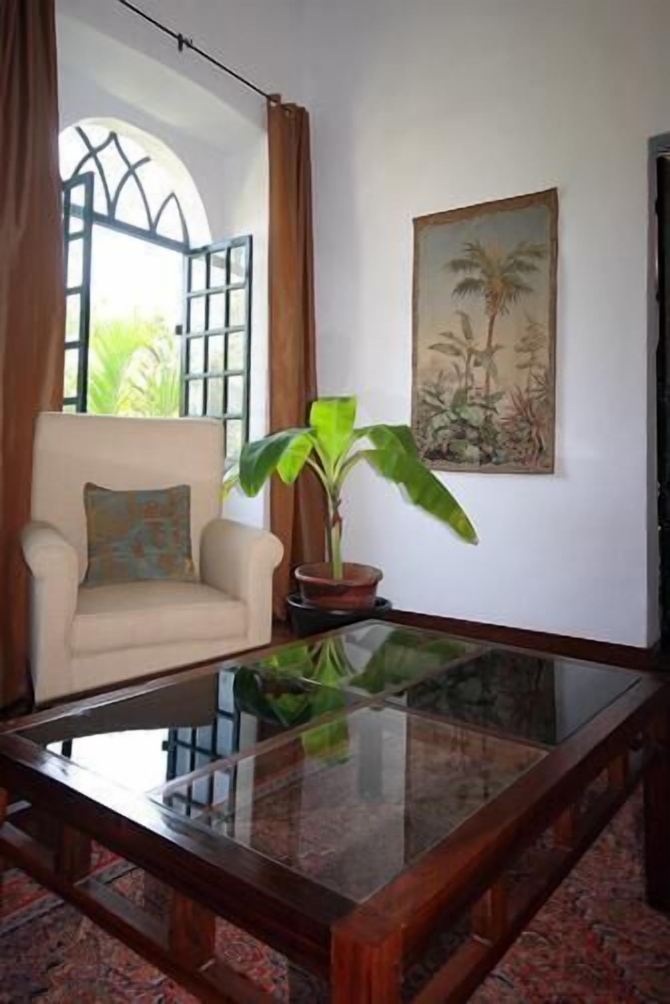 Una habitación con una mesa de centro de vidrio, un sillón color beige, una planta en maceta y un cuadro con plantas tropicales en la pared.