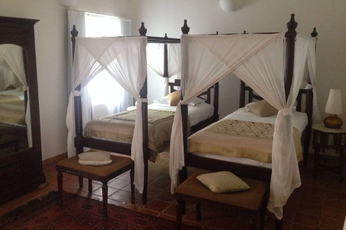 Ein Zimmer mit vier Betten, jedes mit Baldachin. Im Vordergrund steht ein Holztisch und ein Stuhl.