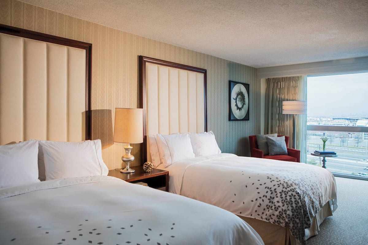 A hotel room with two beds, a red armchair, a side table with a lamp, and a large window offering a city view.