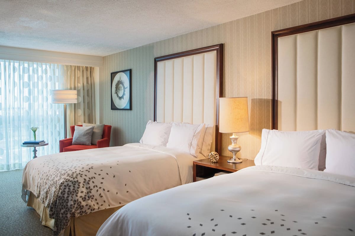 A hotel room with two beds, a red armchair, a small table with a lamp, and a framed picture on the wall.