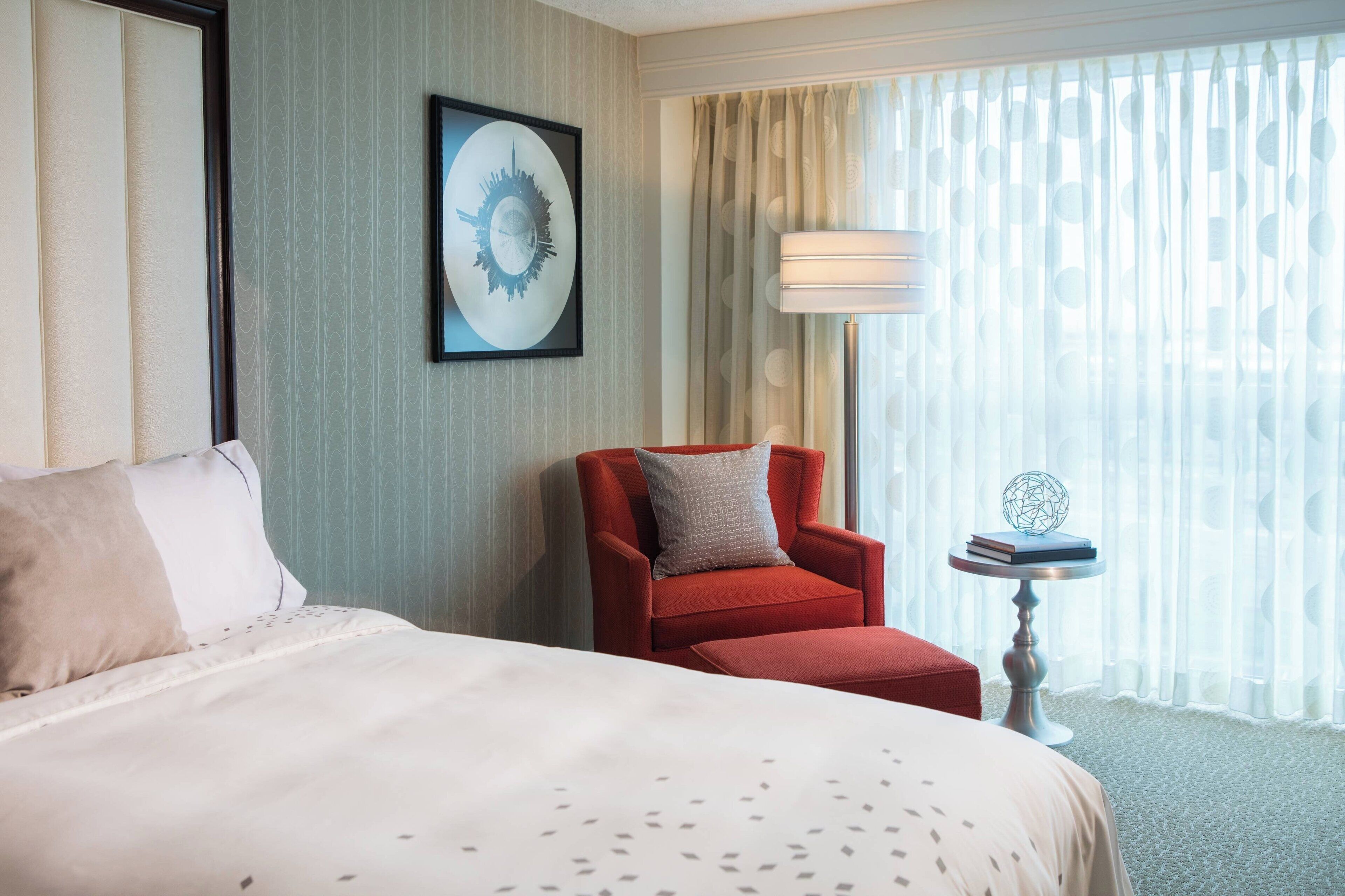 A hotel room with a large bed, a red armchair, a small table with a globe and books, and a window with sheer curtains.