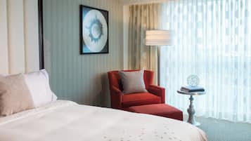 A hotel room with a large bed, a red armchair, a small table with a globe and books, and a window with sheer curtains.