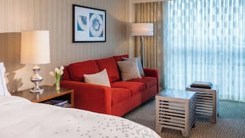 A hotel room with a red sofa, a bedside table with a lamp, a small wooden stool, and a large window with sheer curtains.