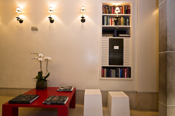 Un área de recepción con una mesa roja, estanterías, lámparas de pared y una orquídea en maceta.