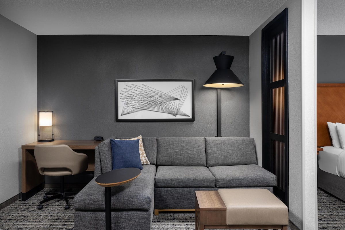 Une chambre d’hôtel moderne avec un canapé gris, une petite table ronde, un bureau avec une chaise, une lampe et une œuvre d’art abstraite encadrée accrochée au mur.
