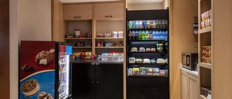 Quarto pequeno e bem organizado, com geladeira, prateleiras cheias de lanches e bebidas, e micro-ondas em uma bancada.