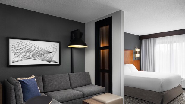 A modern hotel room with a grey sofa, a small wooden table, a bed with white linens, a black floor lamp, and a framed abstract artwork on the wall.