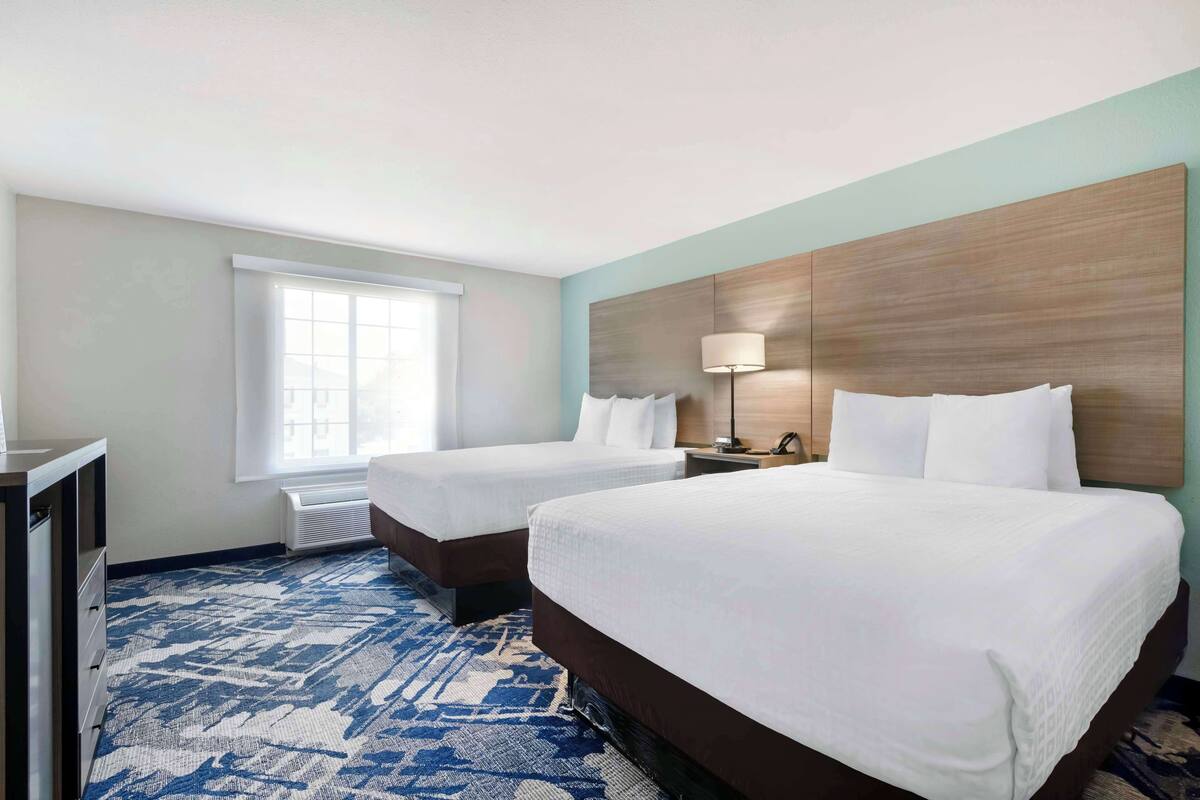 A hotel room with two beds, a desk, a lamp, and a window.
