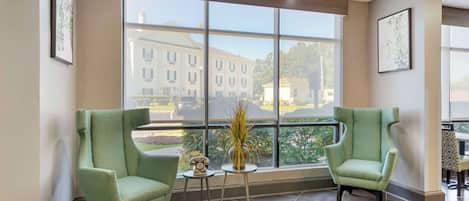 A room with two green armchairs, a small table with a telephone and a vase with plants, and a large window offering a view of buildings and trees.