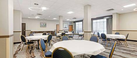 Un salón de conferencias de hotel con varias mesas y sillas, piso alfombrado y un ventanal con cortinas.