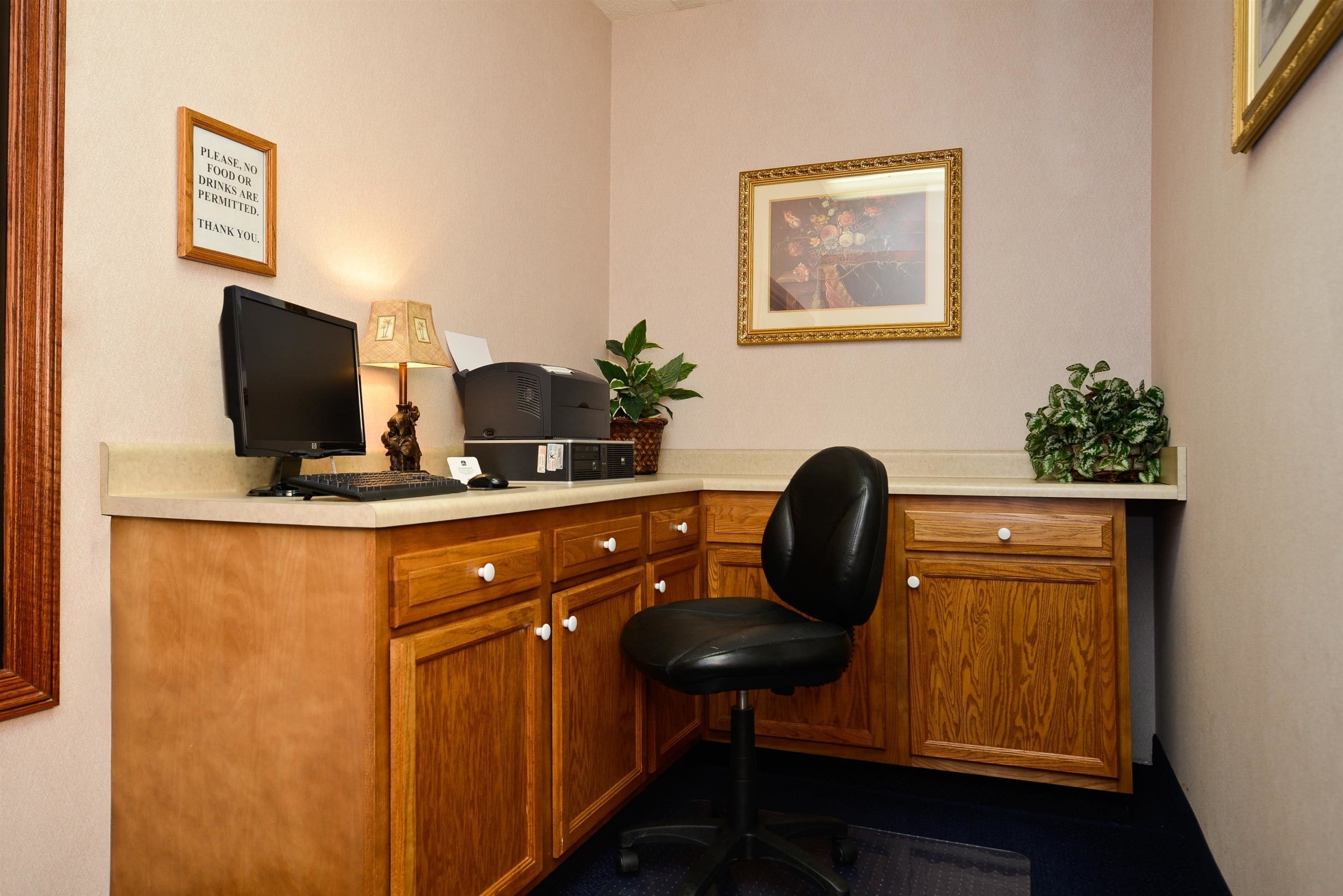 A small office with a desk, computer, keyboard, and a chair. There is a framed picture on the wall and a sign above the desk.