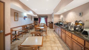 Un comedor de hotel con mesas y sillas de madera, un mostrador de bufé con diferentes opciones de comida, y una zona de cocina con armarios y electrodomésticos.