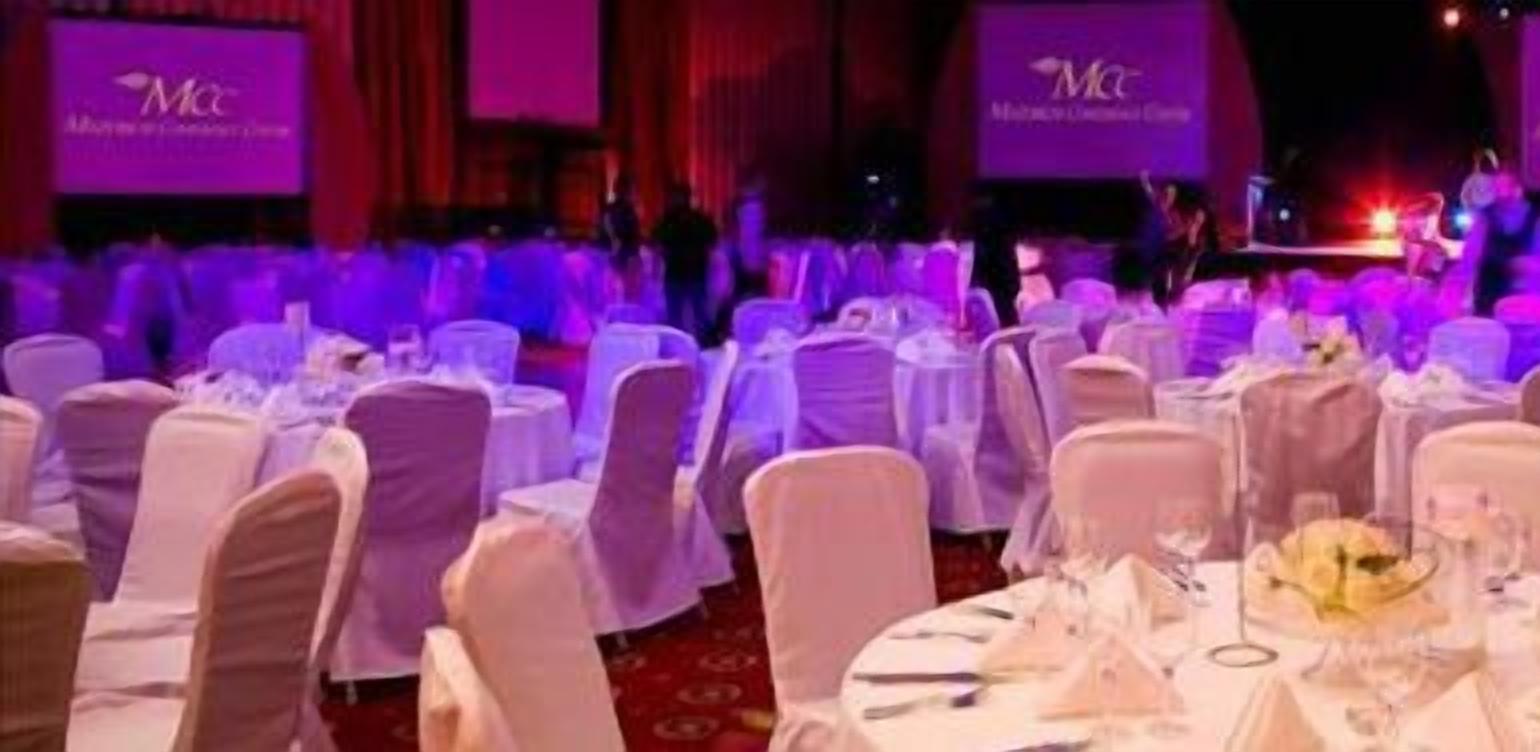 A banquet hall with round tables set for a formal event, featuring a stage with a large screen displaying the event's name.